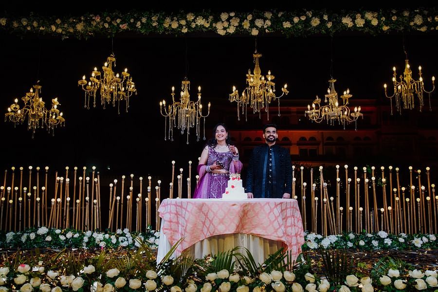 Bride and groom standing behind a decorated table under warm golden lights and chandeliers at a nighttime wedding reception.