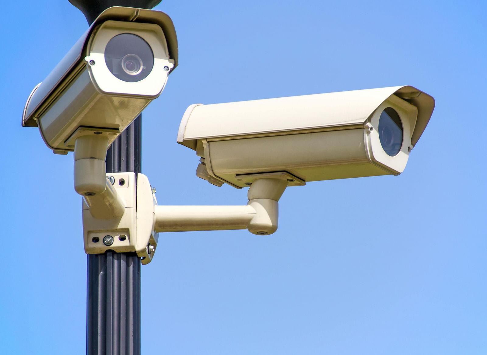 Two white CCTV security cameras mounted on a grey pole against a clear blue sky.