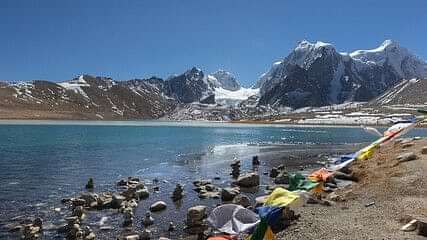 A scenic view of Gurudongmar Lake in Sikkim, with snow-capped mountains in the background and colorful prayer flags along the shoreline.