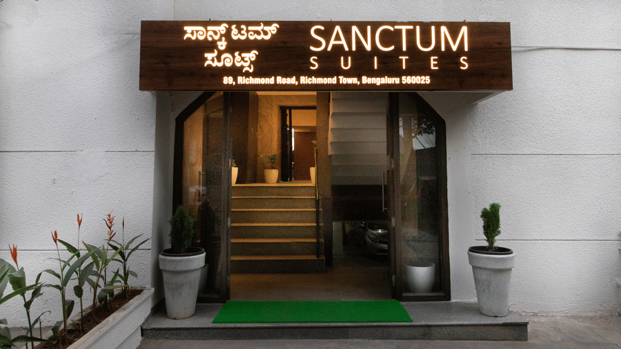 The front entrance of Sanctum Suites, Richmond Road, Bangalore features a dark wooden sign above glass doors, white walls, potted plants, and a green mat.
