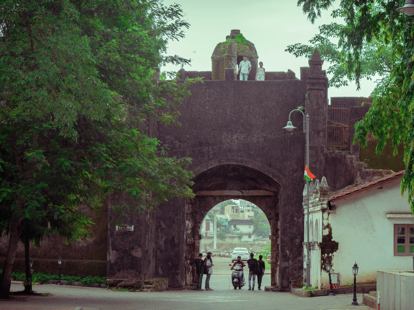 A dark stone fort with a large central arch, surrounded by green trees, with several people standing near the archway and 2 others visible at the top of the structure.
