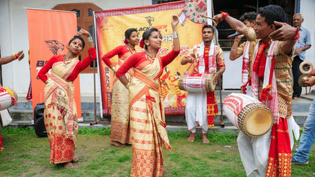 Group of women in cream and red saris dance in a row while two men in white attire play drums on a grass lawn beside an event banner backdrop.