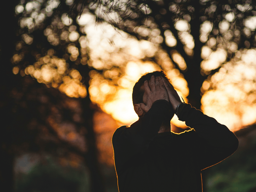 A person silhouetted against a sunset through trees, hands raised to their head