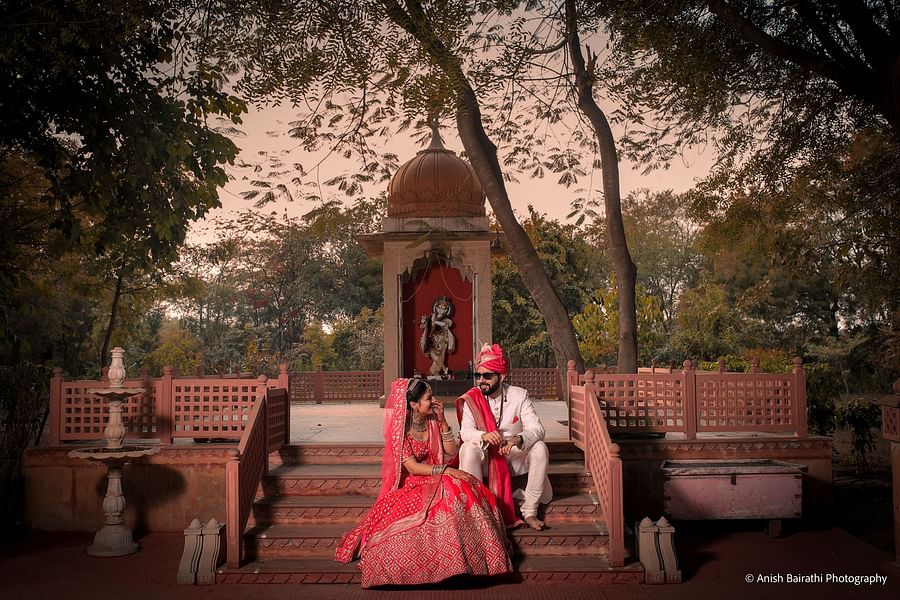 A newlywed couple in wedding attire sits on temple stairs, sharing a joyful moment in a serene outdoor setting.- Umaid Palace - A Lakeside Heritage Palace Resort