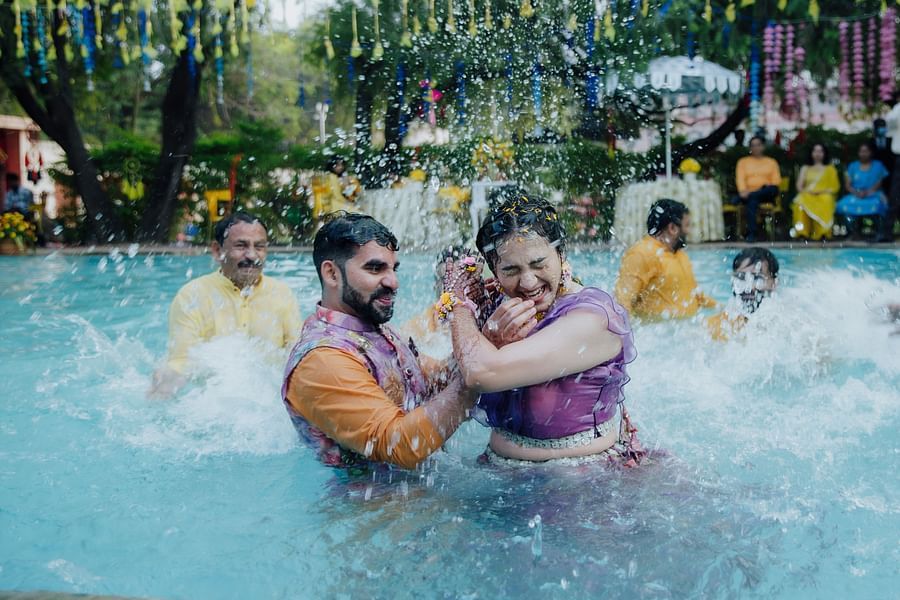 Bride and groom playfully splashing water while dancing together in a pool during a daytime celebration.