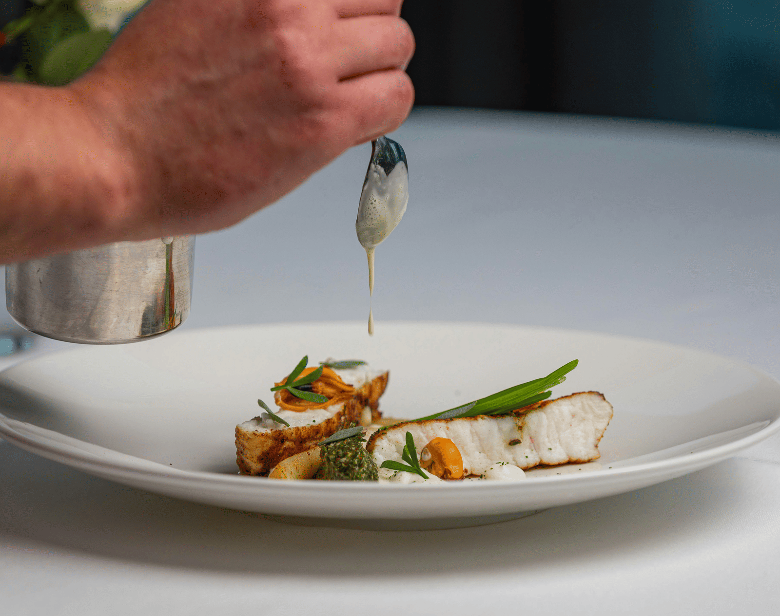 A close-up of a chef's hand carefully pouring a creamy sauce onto a finely plated gourmet meal.