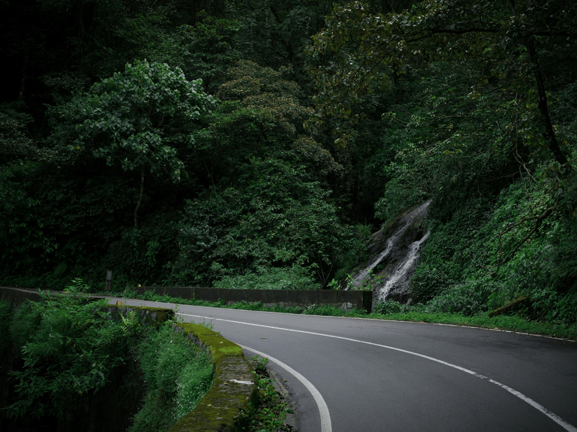 A view of the highway leading to Valparai with a mountain on the side having a small waterfall and trees.