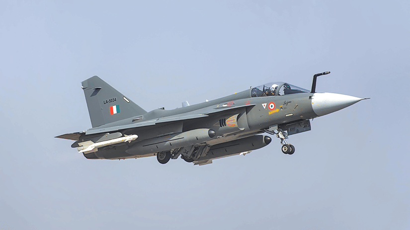 An Indian Air Force HAL Tejas multirole fighter jet, painted grey, flying low with its landing gear partially extended against a pale sky.