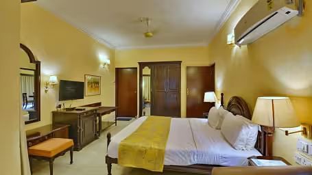  A spacious room with a large bed, a wall mounted television opposite, a dressing table with a chair and wall-mounted mirror beside it, and an air conditioner positioned directly above the bed.