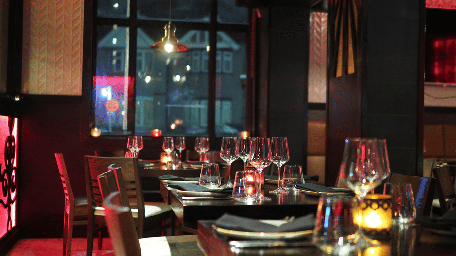 A moody interior shot of a restaurant dining table set with wine glasses and candles, seen through a dark window.