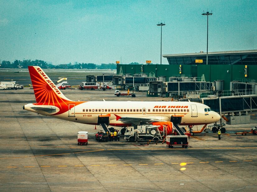 An Air India aeroplane is stationary at an airport terminal, surrounded by ground support vehicles and personnel on the tarmac.