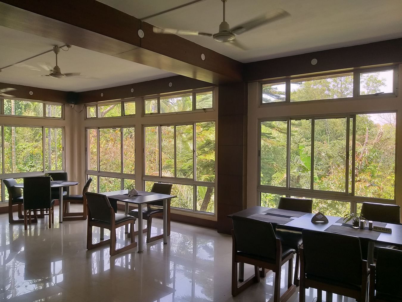 A bright and airy hotel dining space with tables set for service, surrounded by large windows offering views of greenery.