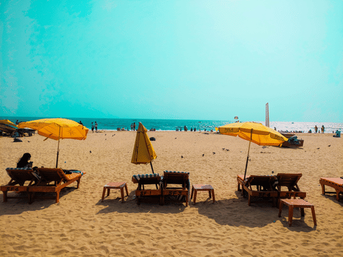 A sandy beach featuring several wooden lounge chairs arranged in rows under open yellow umbrellas, with distant figures visible near the ocean water line.