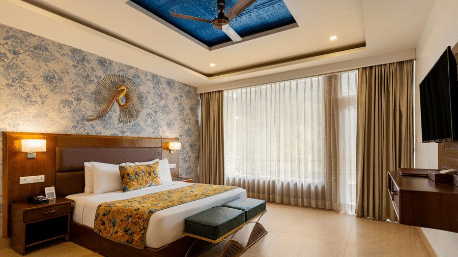 A spacious bedroom featuring a neatly made bed, 2 side cabinets, a wall with blue wallpaper, a decorative ceiling, and sheer and opaque curtains | Summit by the Ganges Beach Resort & Spa, Rishikesh