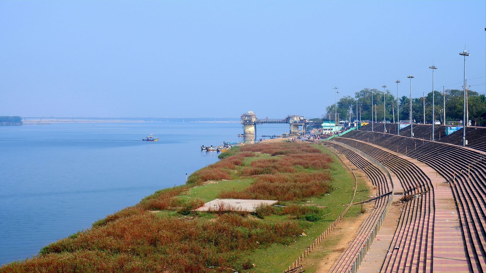 A tranquil riverside scene with a paved walkway, lush greenery, and a distant watchtower under a clear blue sky.