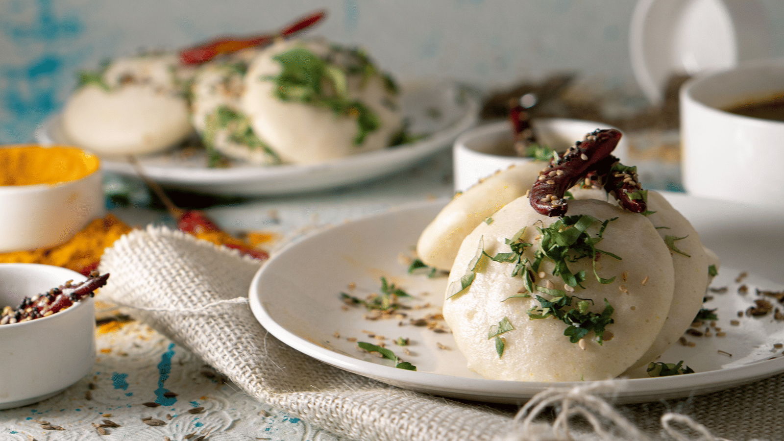 Soft steamed rice idlis garnished with chopped herbs and dried red chilies served on white ceramic plates with spices in the background.