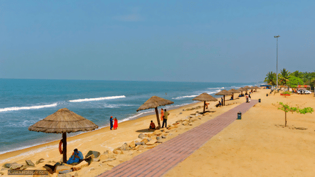 A sandy beach with ocean waves, a paved walkway along the shore, and shade umbrellas placed at regular intervals.