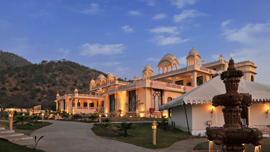 The facade of Rajasthali Resort & Spa, Jaipur, glows against the evening sky, with hills in the background.