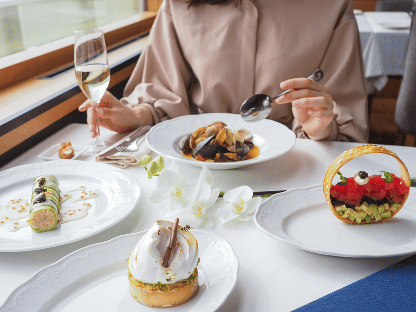 A woman dining at a restaurant table with multiple white plates of gourmet desserts and a main course dish alongside a wine glass and fresh white flowers near a bright window.