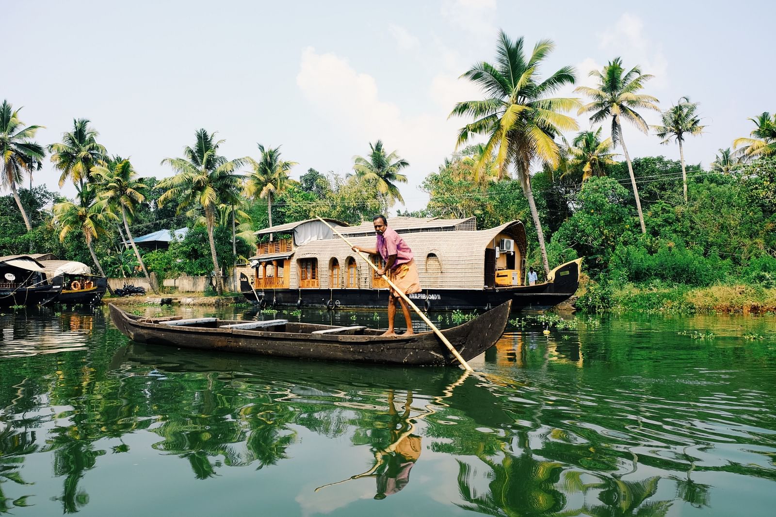 A houseboat with a thatched roof gently navigates the serene backwaters, framed by tropical palm trees under a clear sky- Kumarakom