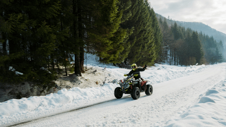 An adventurer riding an all-terrain vehicle (ATV) through a snowy path in a pine forest.