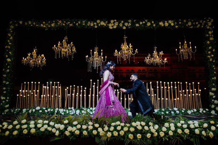 Bride and groom on a stage bowing toward each other with warm lights and elegant floral decor in the background.