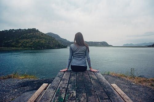 A woman sitting a the edge of a large wooden piece, facing a water body, and green hills on a cloudy day.