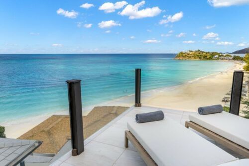 Beach loungers are set up on a deck overlooking a calm, blue ocean with a sandy beach in the background.
