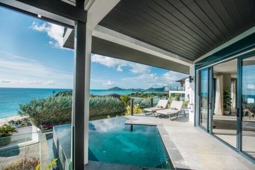 A modern villa features an infinity pool overlooking a vast expanse of the blue ocean under a clear sky.