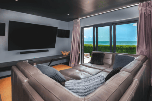A modern living room features a large curved sectional sofa facing a wall-mounted television, with expansive windows offering a view of the ocean.