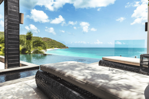 A comfortable outdoor lounge area with cushions overlooks a breathtaking view of the ocean and a distant island under a blue sky.