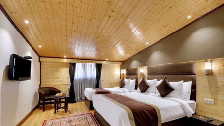 A premium triple room in Gangtok at the Sumitel Shangrila Deorali, with a twin and a double bed opposite a television, along with a chair, a table, and a window.