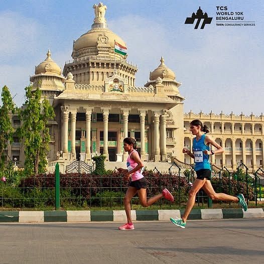 Two female runners participating in the TCS World 10K Bengaluru race, running past the iconic Vidhana Soudha building.