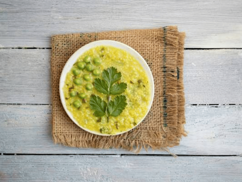 A bowl of cooked dalia with green peas and coriander leaves on a jute mat, on a white wooden surface, seen from above