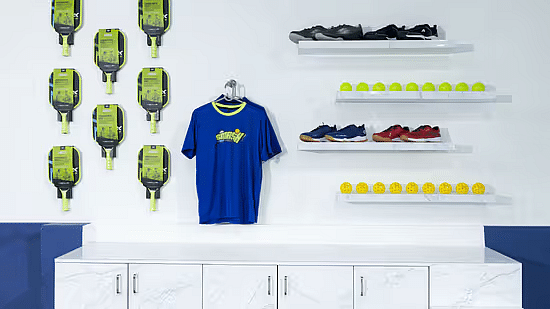 A white wall on which various sportswear items have been placed including a jersey, a shoe rack and two shelves full of balls above a table at Jenneys Residency in Coimbatore.