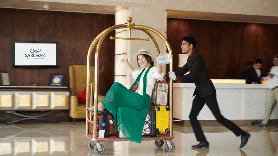 A bell boy pushing a bell trolley with a woman on top of it.