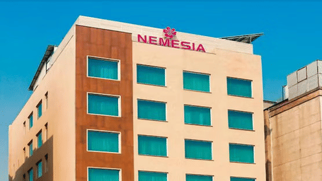 Daytime view of the Nemesia hotel building facade with teal-tinted windows, wooden accents, and palm trees in front.