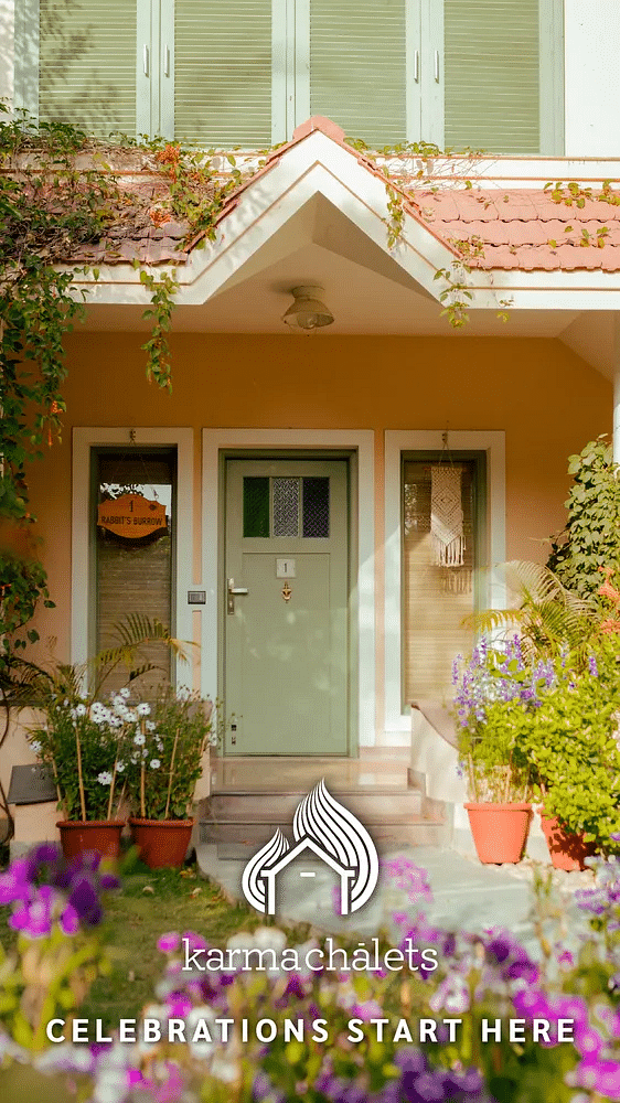 Quaint house entrance with a pale green door, flowerpots, and a red roof, framed by lush garden blooms at Karma Chalets.