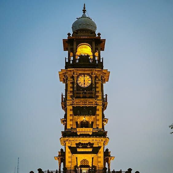 The clock tower at Jodhpur illuminated with lights set against a clear background.