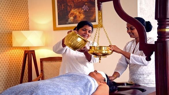 A person receiving a treatment on their face with liquid being poured from a golden container, indoor setting - Shreyas Retreat