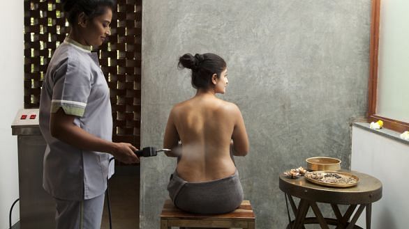 A person receiving a treatment on their back, with another person standing nearby, indoor setting - Shreyas Retreat