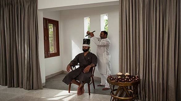 A person seated in a chair, wearing a dark garment with a white headdress, in a room with a window - Shreyas Retreat