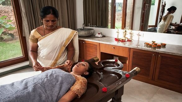 A person receiving a massage on a table, with another person standing nearby, indoor setting - Shreyas Retreat