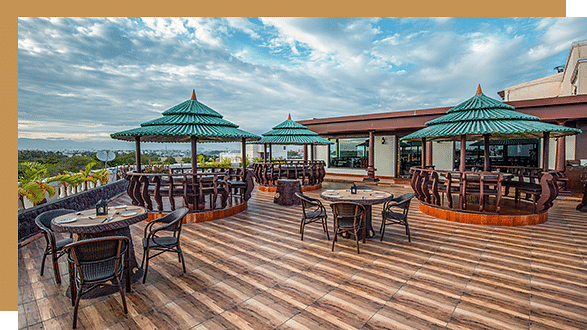 A spacious rooftop Kayaloram terrace features wooden decking, round tables with chairs, and shaded seating areas with green, gazebo-style roofs. The scene is set under a partly cloudy sky.