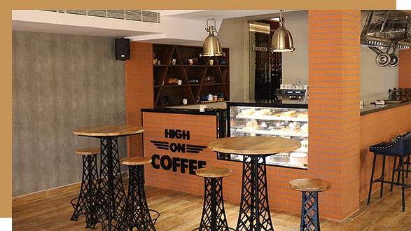A High On Coffee dining at Jenneys Residency interior with wooden tables, industrial-style stools, a display case with pastries, and a counter that reads HIGH ON COFFEE. Warm lighting and shelves in the background
