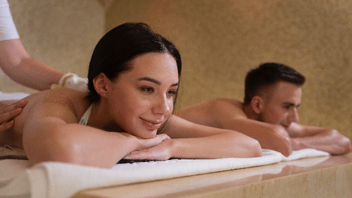 A couple enjoys a relaxing spa experience, lying side by side on massage tables with peaceful expressions in a calming wellness setting