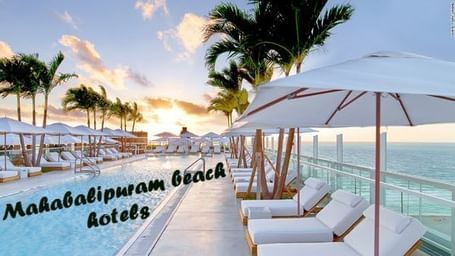 A beachside lounge area at Hotel Mamallaa Heritage, Mahabalipuram, with a pool, multiple white sunbeds, umbrellas, and palm trees with text ‘Mahabalipuram Beach Hotels’ in the image.