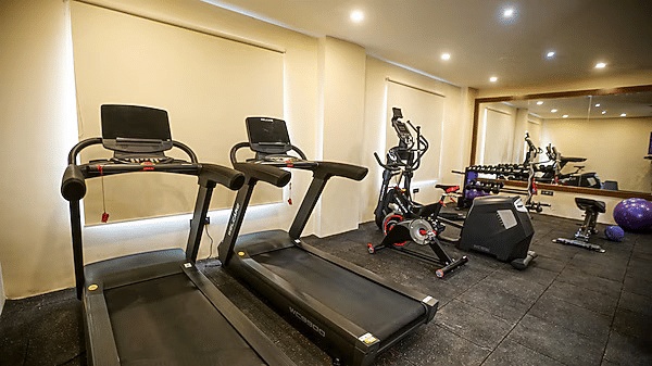 A wide-angle shot of the gym at Leroy Grand, Yeshwanthpur, featuring seral gym equipments, machines, and 2 treadmills with windows in front of them covered with white blinds.