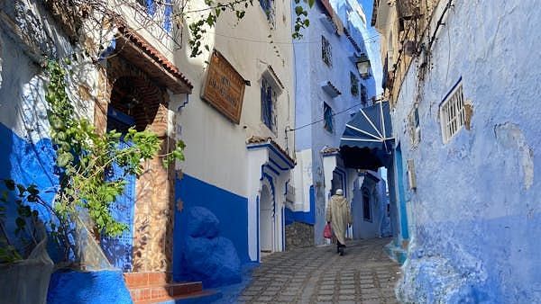An image of a pathway where a woman is seen walking and white and blue buildings on one side and a wall on the other