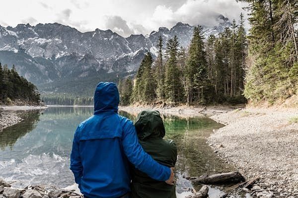 A couple in a valley facing the pine trees, water body, and the majestic mountains.